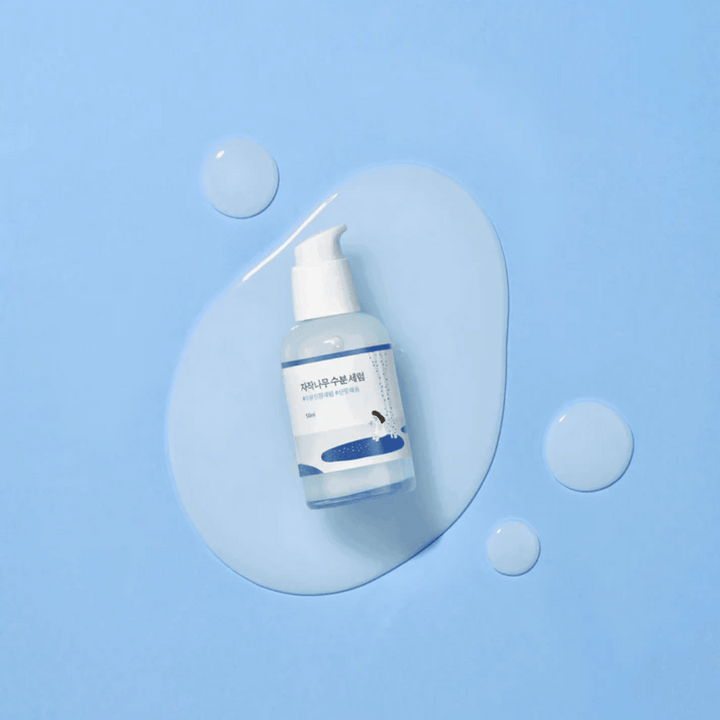 Round Lab Birch Juice Moisturizing Serum i genomskinlig pumpflaska placerad på en blå bakgrund med droppar och utspillt serum.