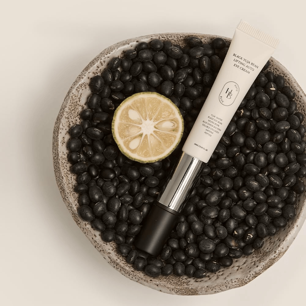 Black Yuja Bean Lifting Auto Eye Cream liggande i en skål fylld med svarta bönor och en halverad citrusfrukt – en visuell representation av produktens naturliga ingredienser.
