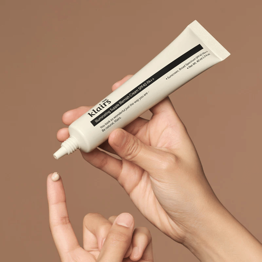 En hand håller en tub av Klairs Illuminating Supple Blemish Cream SPF40 PA++ och trycker ut en liten mängd kräm på ett finger, mot en brun bakgrund.