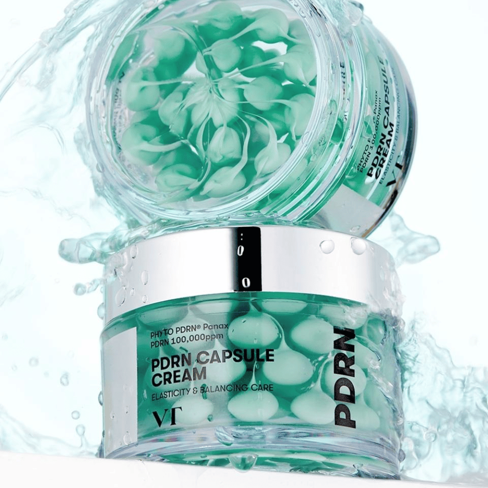Två burkar av VT PDRN Capsule Cream staplade med vattenstänk runtom. De genomskinliga burkarna visar gröna kapslar inuti, med texten "Elasticity & Balancing Care" synlig på etiketten.