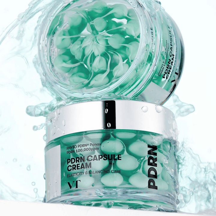 Två burkar av VT PDRN Capsule Cream staplade med vattenstänk runtom. De genomskinliga burkarna visar gröna kapslar inuti, med texten "Elasticity & Balancing Care" synlig på etiketten.
