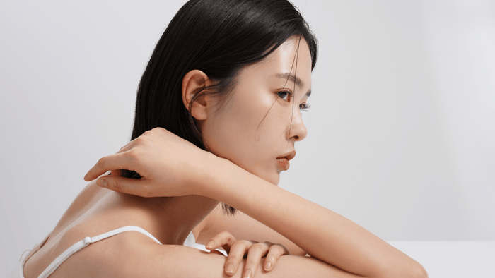 Närbild av en kvinna med slät och strålande hud, sittandes i en avslappnad pose. Hennes naturliga skönhet och minimalistiska bakgrund framhäver en känsla av renhet och koreansk hudvårdsestetik.