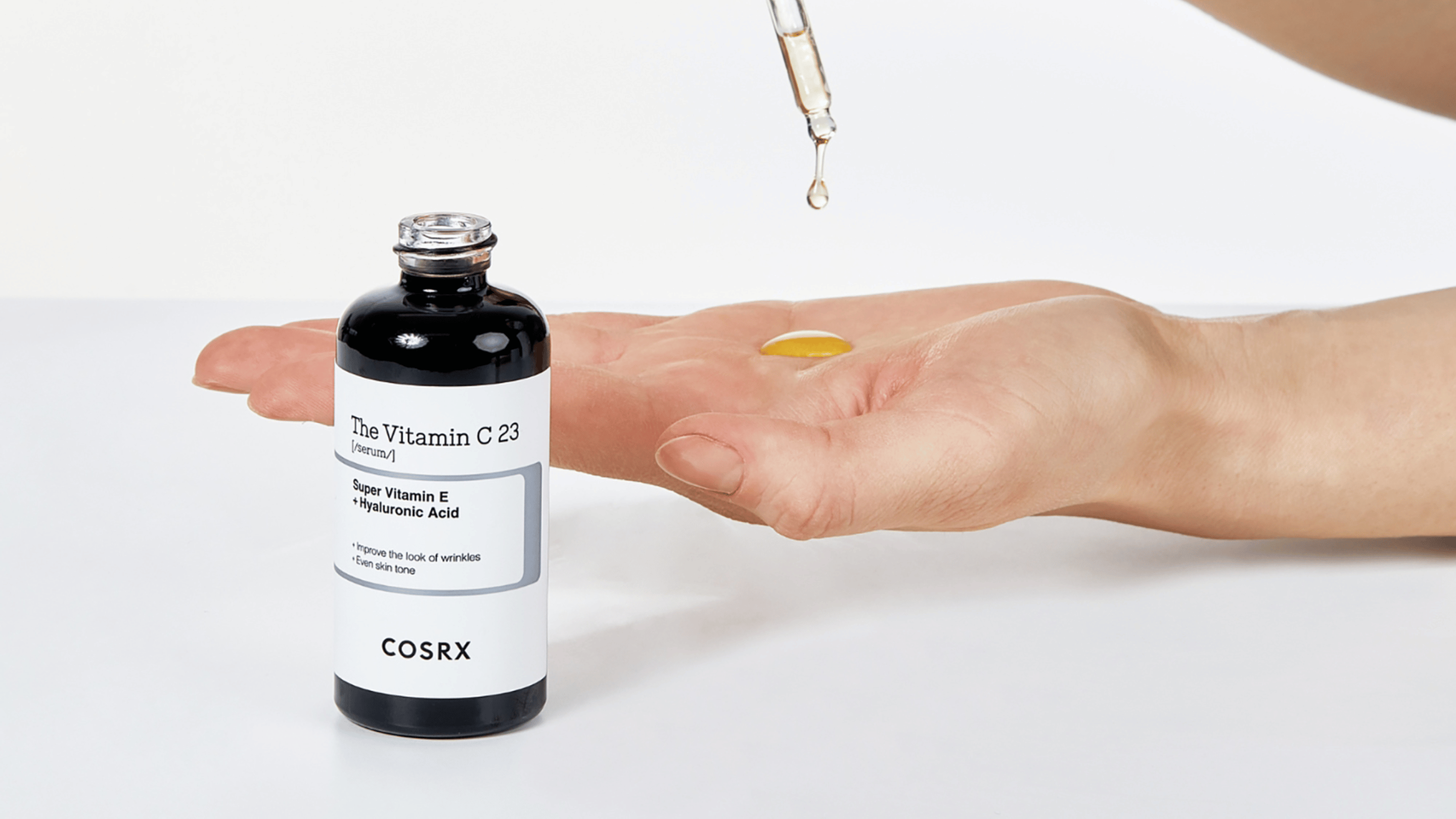 COSRX The Vitamin C 23 Serum med Super Vitamin E och hyaluronsyra. En hand håller serumet, och en pipett droppar gul vätska i handflatan. Produkten är designad för att reducera rynkor och jämna ut hudtonen, perfekt för en strålande hud.