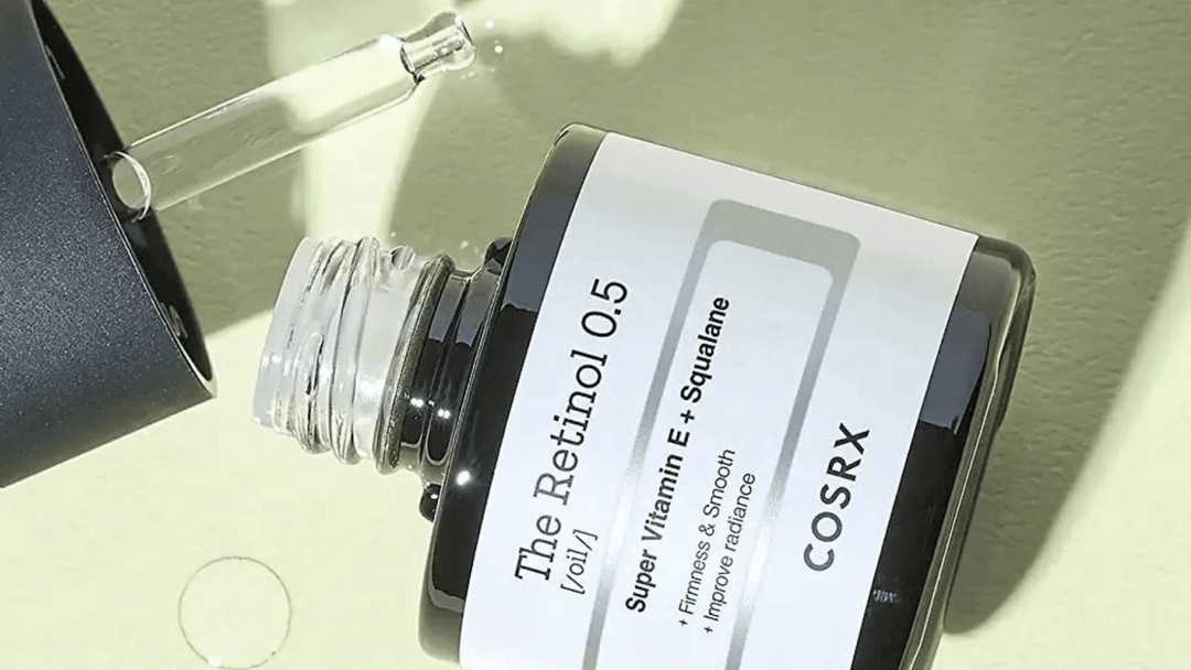 En droppflaska med COSRX The Retinol 0.5 olja ligger på sidan med pipetten droppande ut produkt, i solljus mot en grön bakgrund.