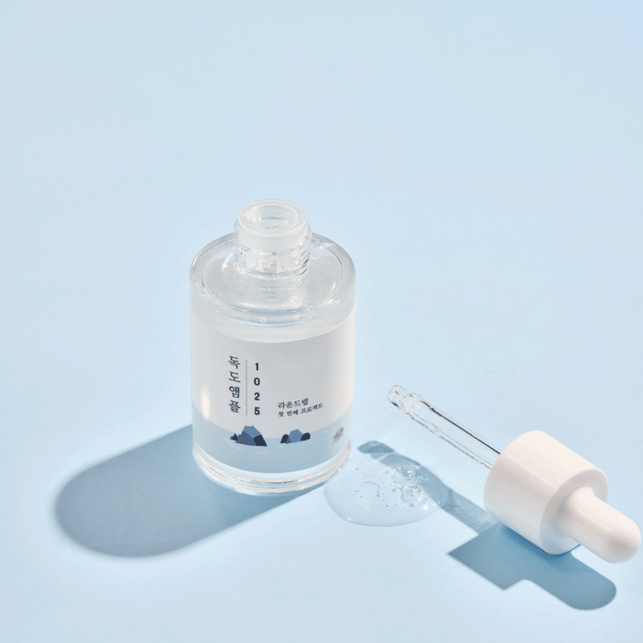 En öppen flaska av 1025 Dokdo Ampoule med en droppare bredvid, mot en blå bakgrund som skapar en mjuk skugga.