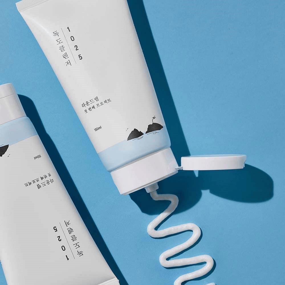 Två tuber av 1025 Dokdo Cleanser på blå bakgrund, varav en med öppen kork och en vågig sträng rengöring utpressad.