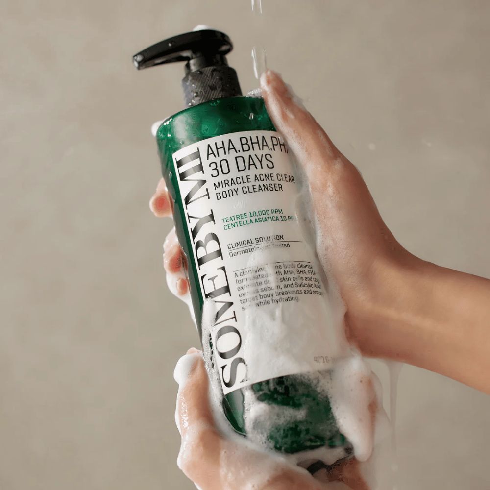 En person håller i en flaska SOME BY MI AHA.BHA.PHA 30 Days Miracle Acne Clear Body Cleanser. Flaskan är grön med en vit etikett och är täckt av lödder från rengöringsmedlet. Personen tvättar sig med produkten medan vatten rinner över händerna.