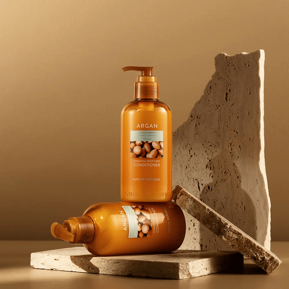 Två flaskor av Nature Republic Argan Essential Moisture Repairing Conditioner på stenunderlag i varm ton.
