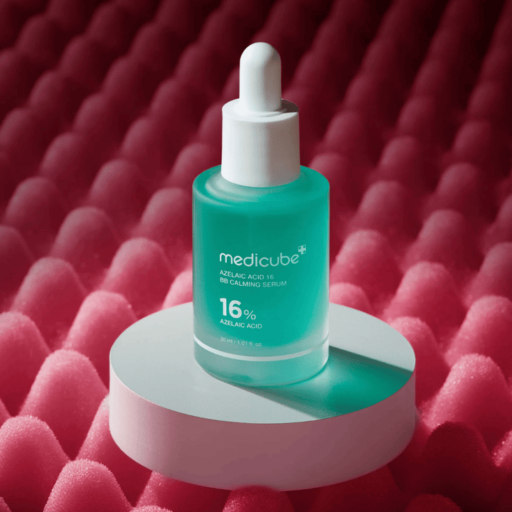 Produktbild av Medicube Azelaic Acid 16 BB Calming Serum placerad på vit podiumyta med rosa strukturerad bakgrund som framhäver serumets lugnande och hudförbättrande profil.