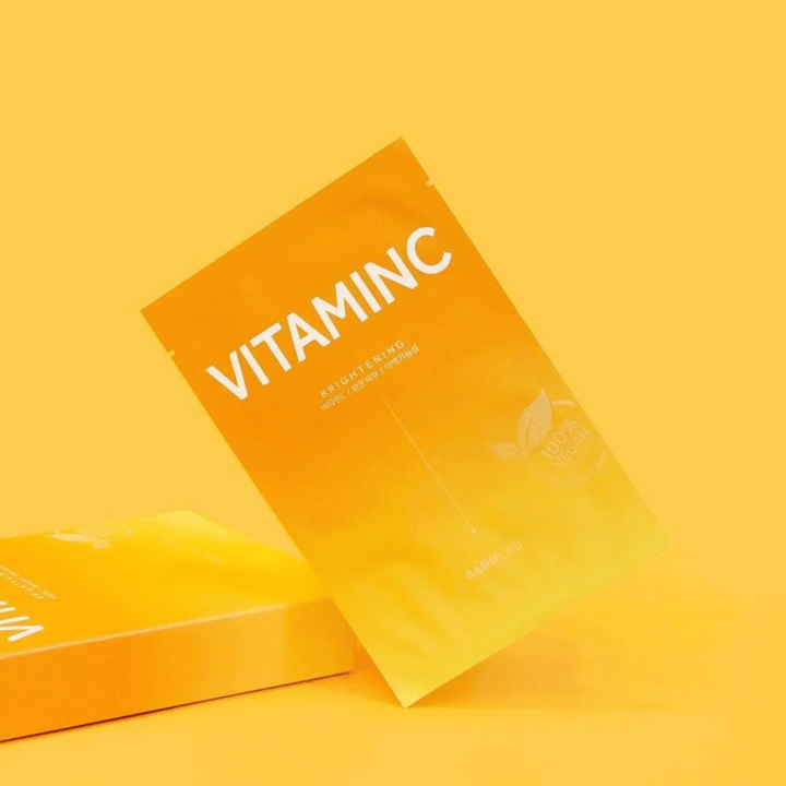 En hög av förpackningar av BARULAB "VITAMINC Brightening" masker, alla märkta som "100% Vegan Product". Förpackningarna har en färgton i olika nyanser av orange och gult, placerade mot en enhetlig gul bakgrund.