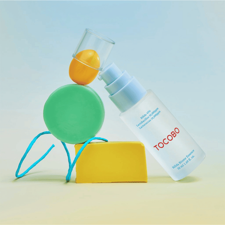 TOCOBO Bifida Biome Essence i frostad pumpflaska med röd logotyp, placerad mot färgglad bakgrund med gröna, gula och orangea geometriska former. Text på flaskan anger Bifida 5 %, Lactobacillus 3 000 ppm och Lactococcus 1 000 ppm.