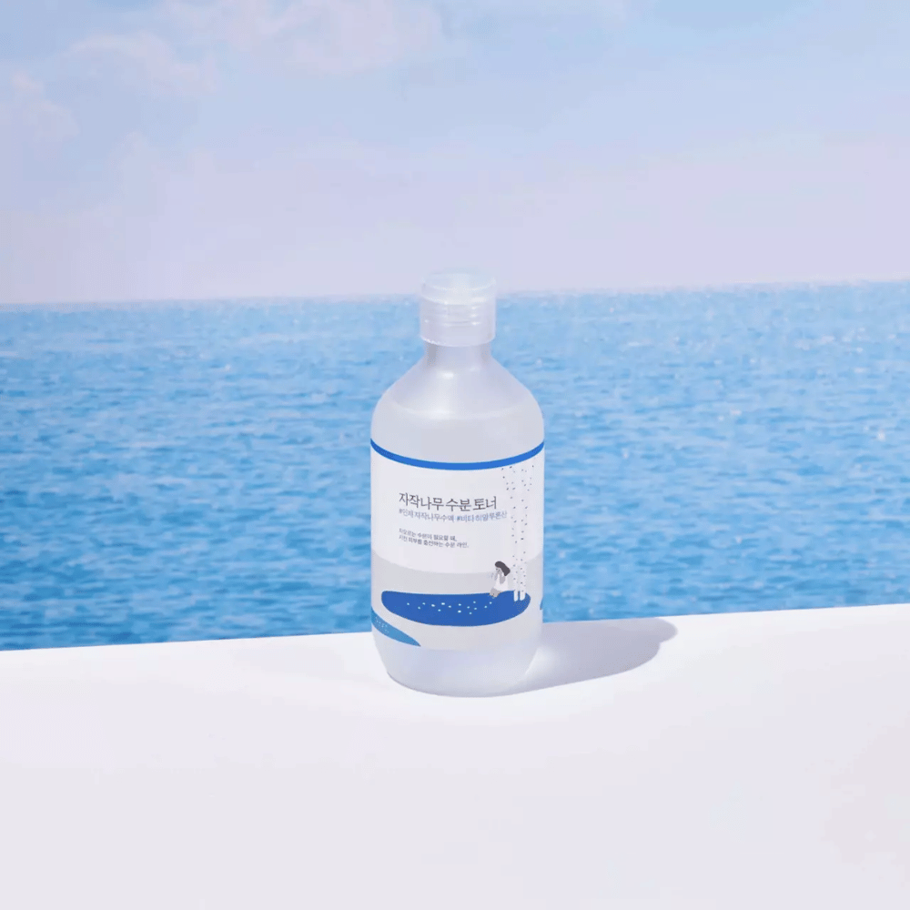Flaska av ROUND LAB Birch Juice Moisturizing Toner står på en vit yta med hav och blå himmel i bakgrunden