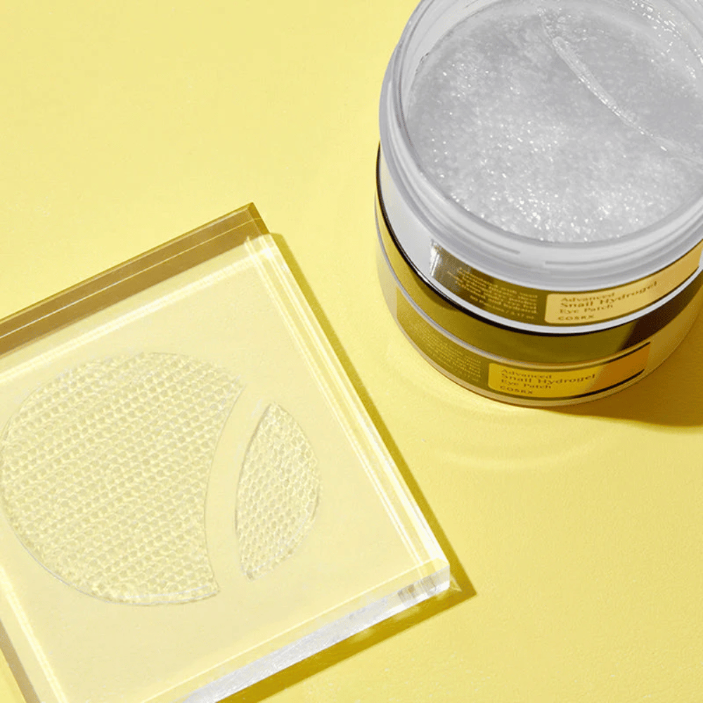 En öppen burk COSRX Advanced Snail Hydrogel Eye Patch står på gul bakgrund bredvid ett genomskinligt underlägg där två ögonpatches ligger utplacerade.