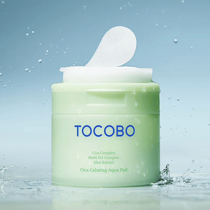 Öppen burk med TOCOBO Cica Calming Aqua Pad där en toner pad sticker upp ur förpackningen. Vattendroppar runt produkten förstärker känslan av intensiv återfuktning och fräschör.