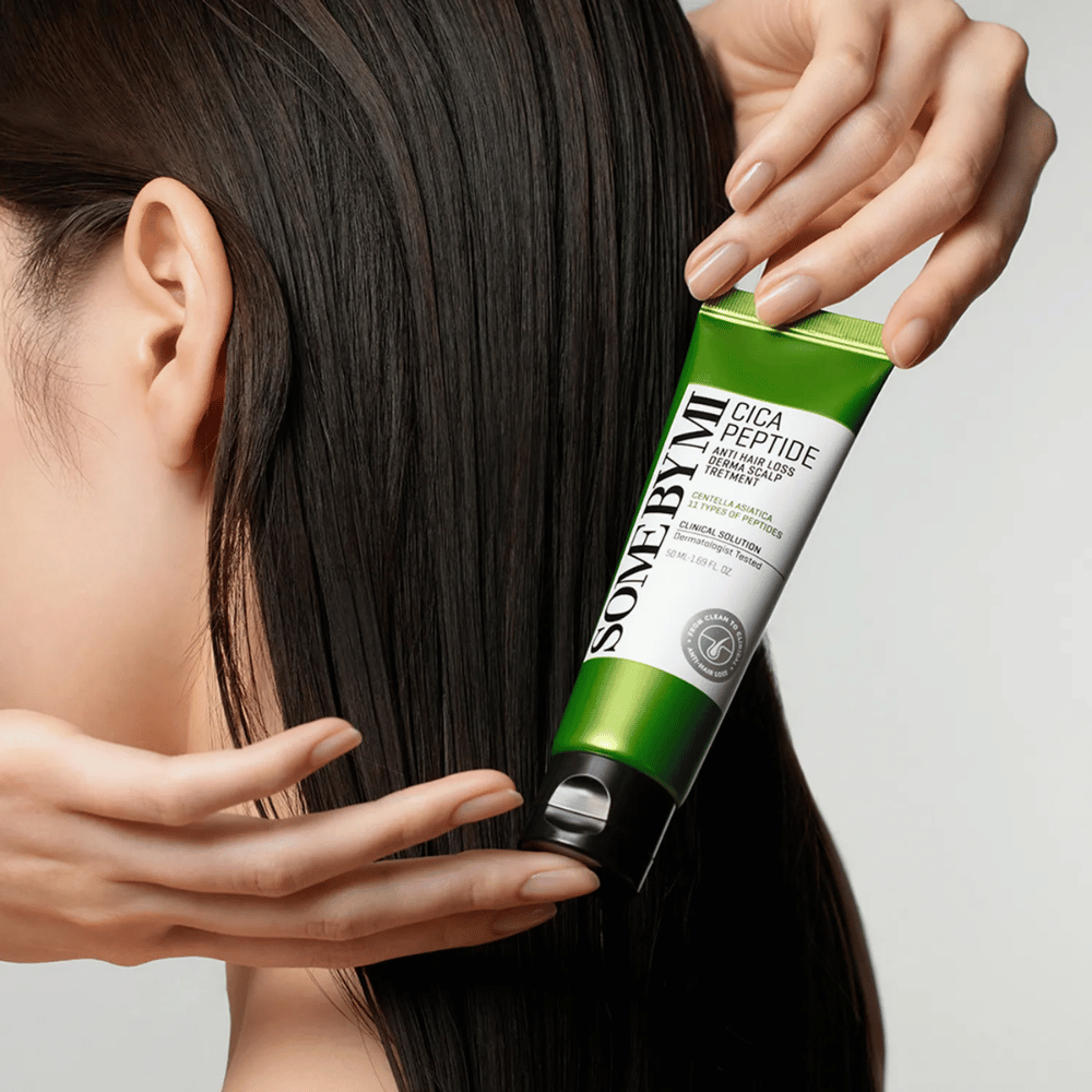Kvinna applicerar SOME BY MI Cica Peptide Anti Hair Loss Derma Scalp Treatment i långt, mörkt hår med en grön och vit tub.