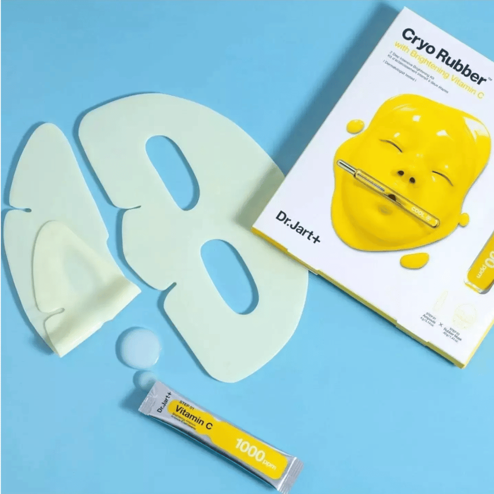 Bild av Dr.Jart+'s 'Cryo Rubber™ med Brightening Vitamin C' skönhetsprodukt. På en blå bakgrund ses en vit gummiansiktsmask, en liten tub märkt 'Vitamin C 1000ppm', och en låda med produktens namn och bild av den gula masken.