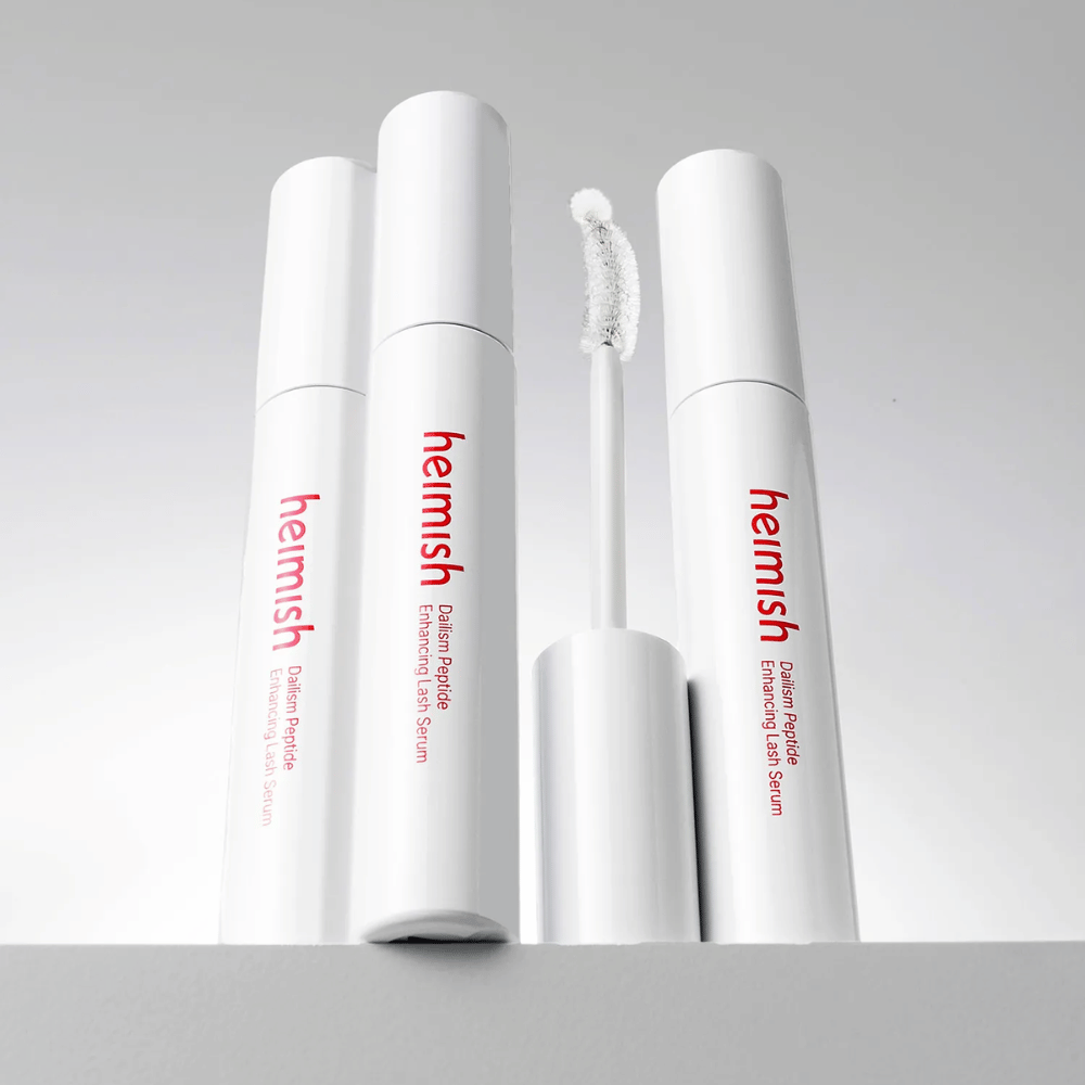 Flera förpackningar av Heimish Dailism Peptide Enhancing Lash Serum visas tillsammans med den böjda applikatorborsten. Bilden framhäver produktens eleganta design och precisionsborste för enkel applicering.