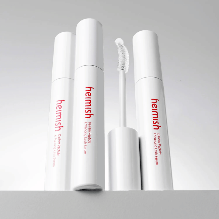 Flera förpackningar av Heimish Dailism Peptide Enhancing Lash Serum visas tillsammans med den böjda applikatorborsten. Bilden framhäver produktens eleganta design och precisionsborste för enkel applicering.