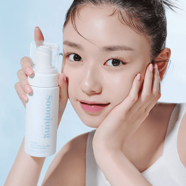 En ung kvinna med en klar hy håller en vit pumpflaska med texten "Soon Jung pH 6.5 Whip Cleanser" från ETUDE. Hon ler mot kameran med en lätt makeup och en fräsch utstrålning mot en ljusblå bakgrund.