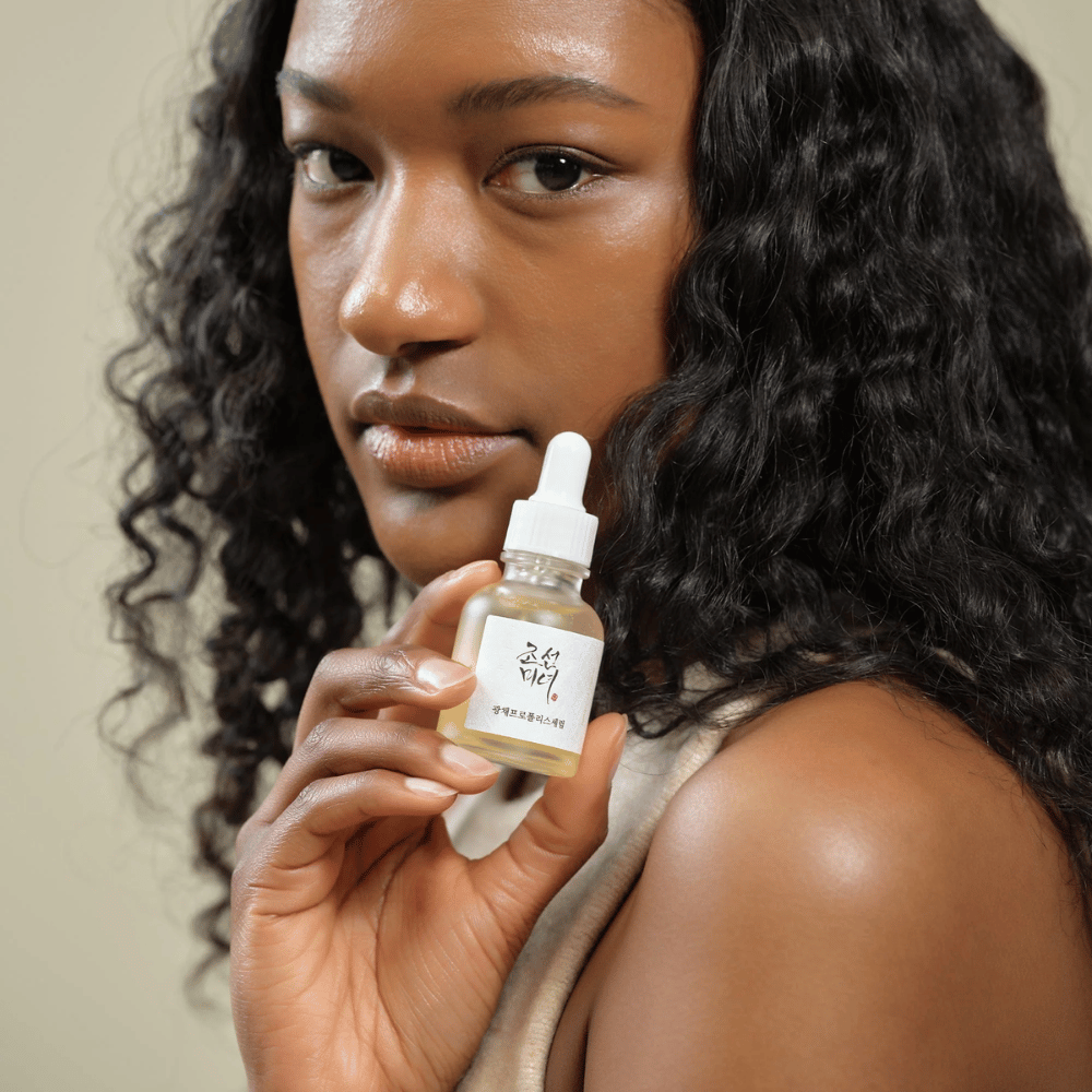 Kvinna med lockigt hår håller upp en flaska Beauty of Joseon Glow Serum med pipett, en återfuktande och lystergivande hudvårdsprodukt.