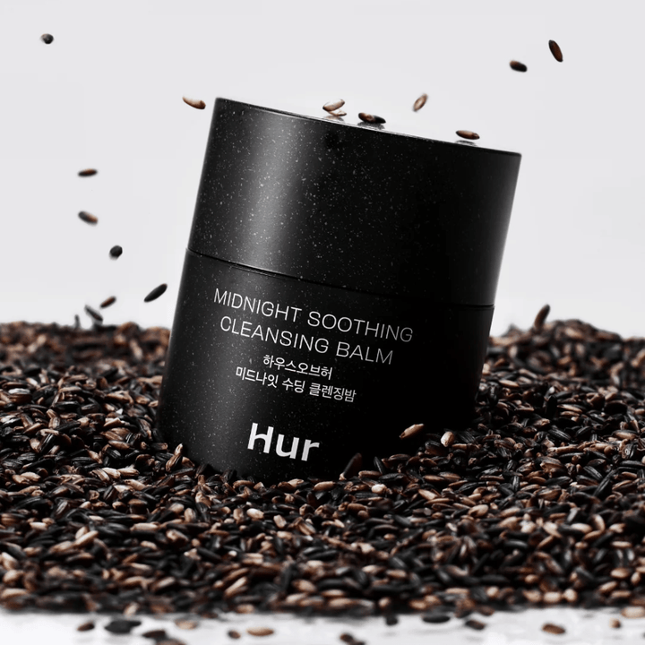Hur Midnight Soothing Cleansing Balm i svart burk placerad på svarta frön. Lug­nande rengöringsbalm som löser makeup och orenheter samtidigt som huden vårdas.
