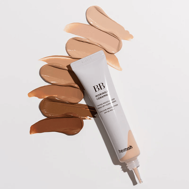 Heimish Moringa Ceramide BB Cream omgiven av flera nyansprover i olika hudtoner. Bilden visar produktens breda färgutbud och den krämiga konsistensen som ger naturlig täckning och jämn hudton.