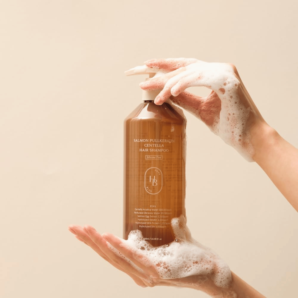 Heveblue Salmon Pullkeratin Centella Hair Shampoo i brun flaska omgiven av skum. Produkten hålls mellan två händer och framhäver en rengörande och vårdande hårvårdsupplevelse med rikligt lödder.