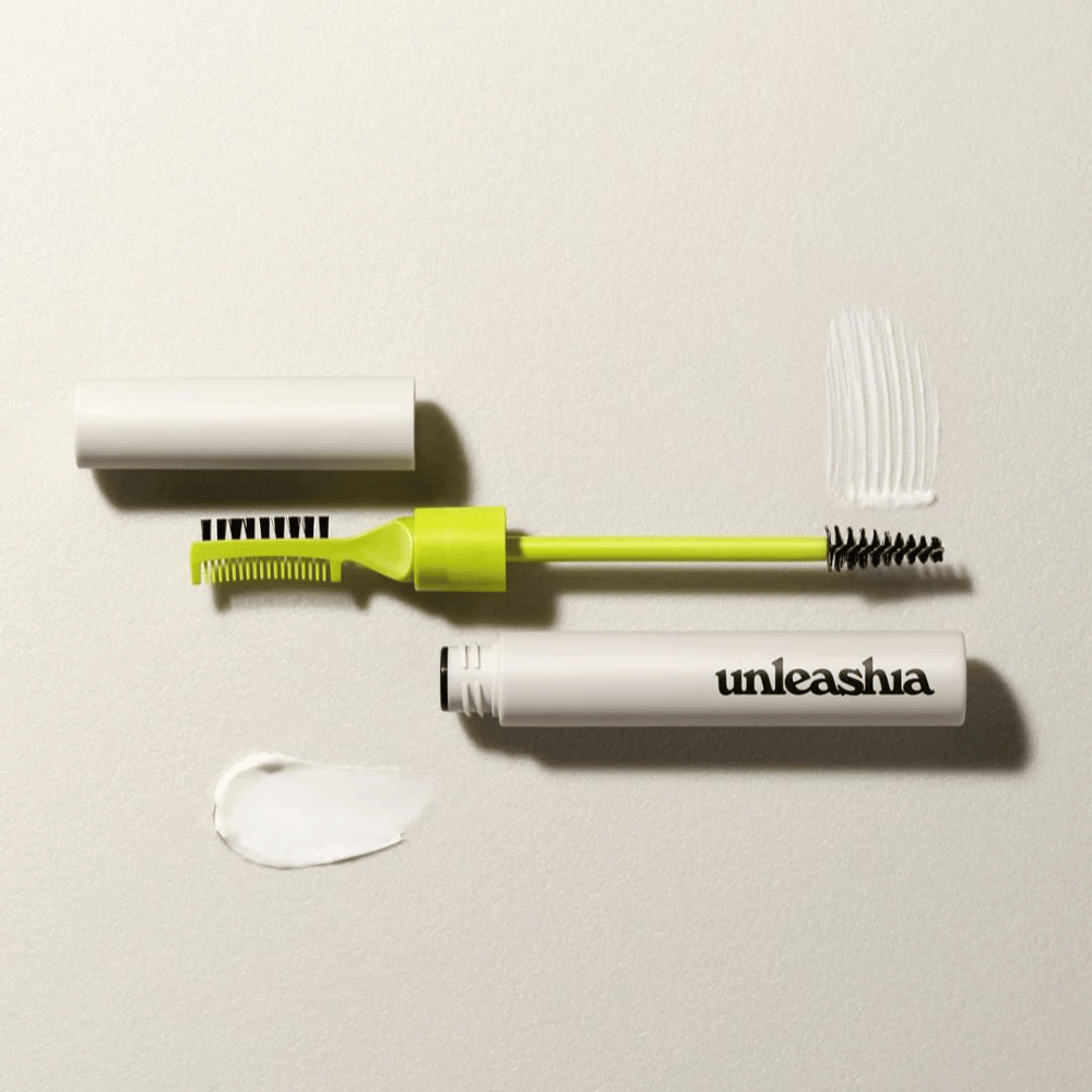 Ett ögonbrynsskulpterande set med en grön kam, svart borste och en vit tub märkt "unleashia" på en ljus bakgrund med en klump av genomskinlig produkt.