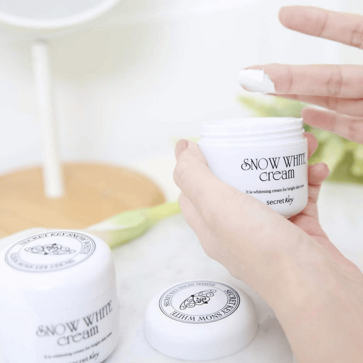 Hand som tar upp vit kräm ur en burk Secret Key Snow White Cream på ett sminkbord.