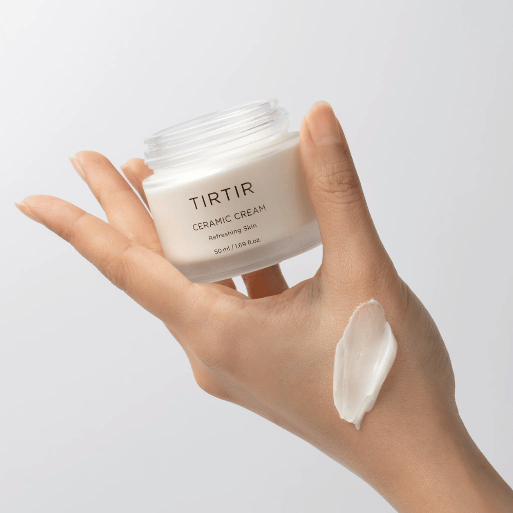 En hand håller en burk med TIRTIR Ceramic Cream. På handens rygg har en mängd av krämen applicerats. Krämen verkar vara tjock och återfuktande. Förpackningen anger att innehållet är för "Refreshing Skin" och mängden är 50 ml / 1.69 fl oz.