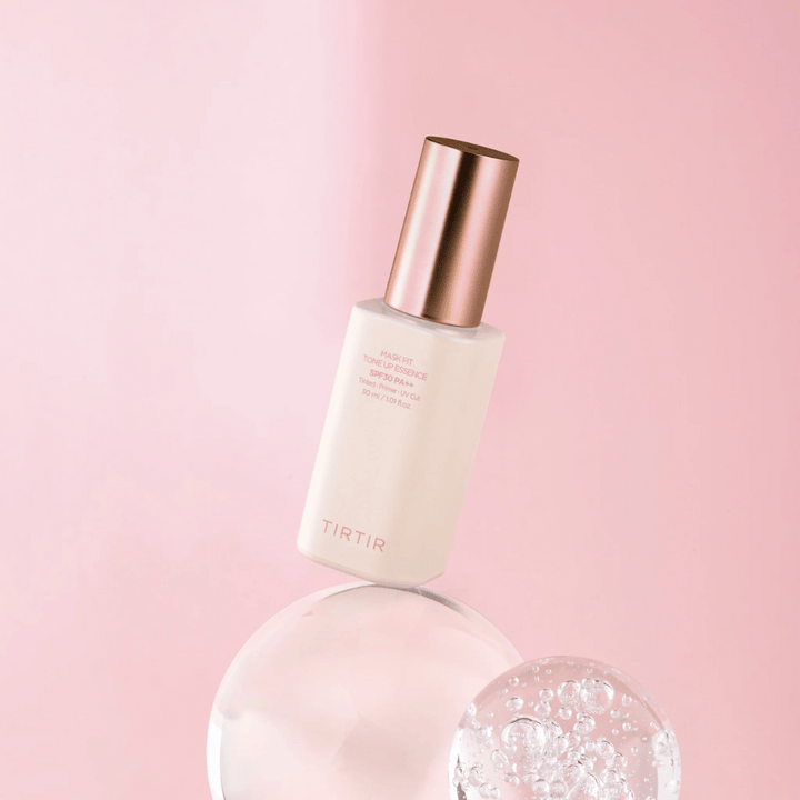 Bilden visar en flaska "TIRTIR MASK FIT TONE UP ESSENCE" med SPF30 PA++ mot en rosa bakgrund, vilket ger en känsla av mjukhet och femininitet.