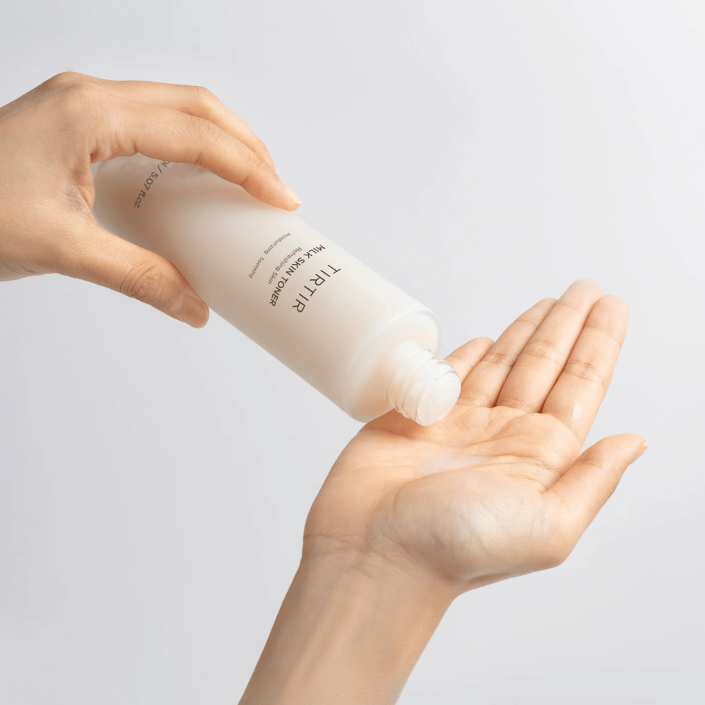 Bilden visar en person som häller ut en mjölkig vätska från en flaska märkt "TIRTIR Milk Skin" på handflatan. Produkten verkar vara en hudtoner eller fuktgivande lotion.