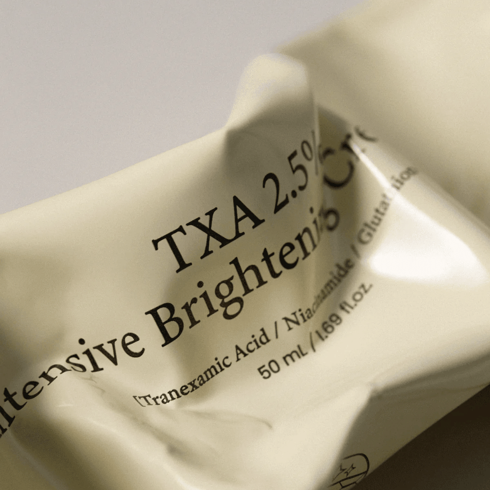 Närbild på AXIS - Y TXA 2.5% Intensive Brightening Cream där den mjuka och minimalistiska tubdesignen står i fokus. Bilden framhäver produktnamn, Tranexamic Acid och Niacinamide samt den eleganta beige förpackningen.