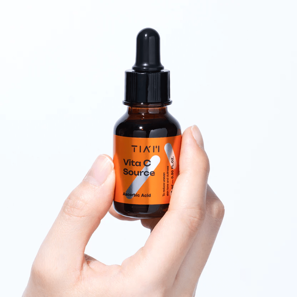 Hand håller i TIA'M Vita C Source, ett C-vitaminserum i brun glasflaska med pipett. Produkten är utformad för att jämna ut hudton och ge lyster.