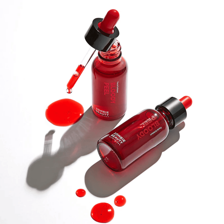 Två röda flaskor med Zombie Beauty Bloody Peel, en stående och en liggande med droppar runtomkring, på en reflekterande yta.