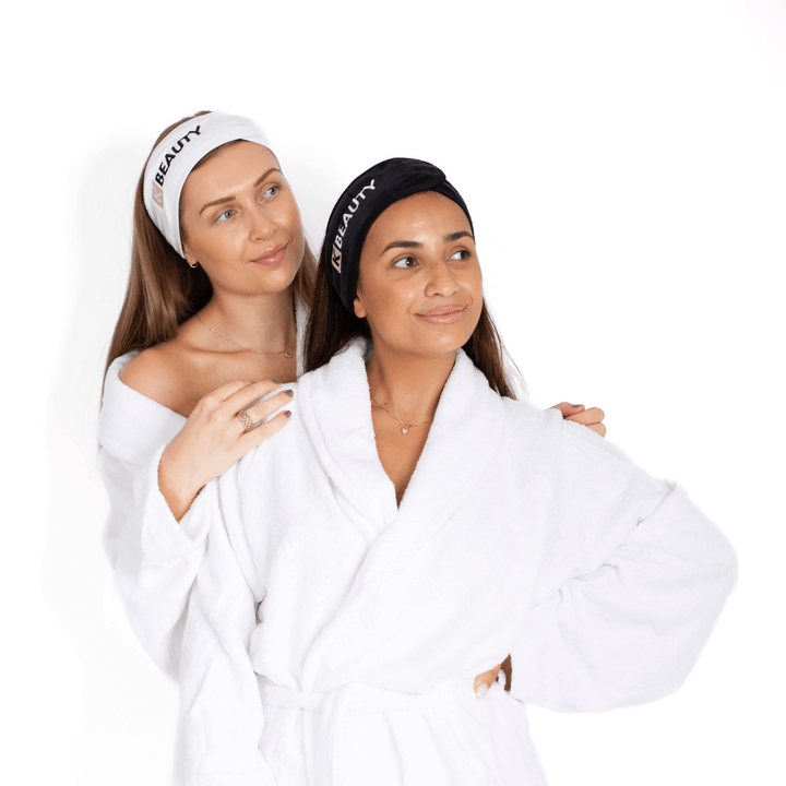 Två kvinnor i vita dräkter poserar för ett foto medan de håller håret ur vägen med K-Beauty Makeup Pannband från K-Beauty.