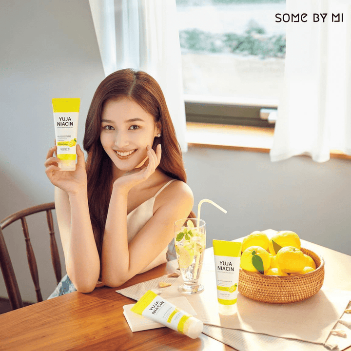 En ung kvinna använder en Yuja Niacin Brightening Peeling Gel 120ml från SOME BY MI för att ta bort döda hudceller vid ett bord med citroner framför sig.